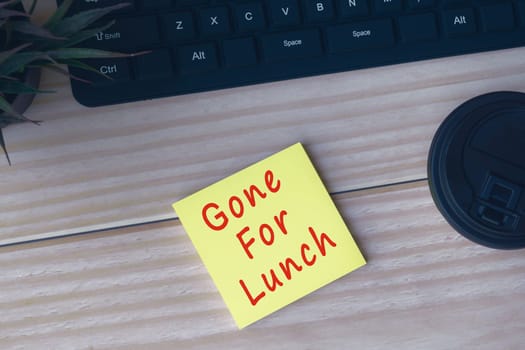 Yellow sticky note with text Gone for Lunch on wooden desk with computer keyboard, potted plan and disposable coffee cup.