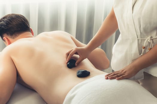 Hot stone massage at spa salon in luxury resort with day light serenity ambient, blissful man customer enjoying spa basalt stone massage glide over body with soothing warmth. Quiescent