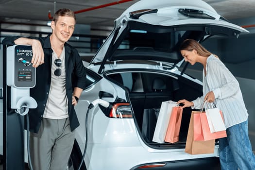 Young couple travel with EV electric car to shopping center parking lot charging in downtown city showing urban sustainability lifestyle by green clean rechargeable energy of electric vehicle innards