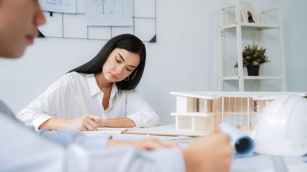 Professional architect engineer drawing blueprint and working together with young beautiful caucasian coworker on meeting table with safety helmet, house model, blueprint scatter around. Immaculate.