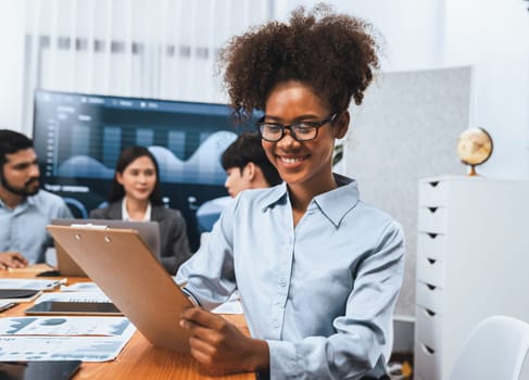 Happy young african businesswoman wearing glasses portrait with group of office worker on meeting with screen display business dashboard in background. Confident office lady at team meeting. Concord