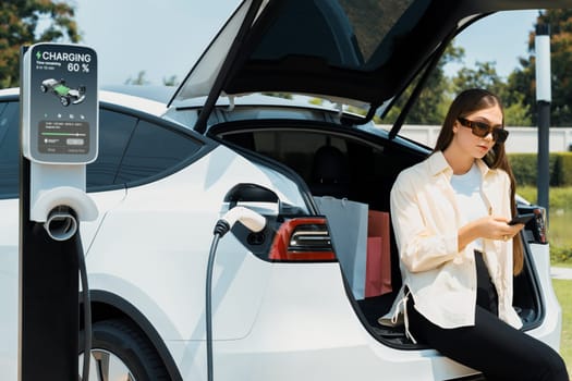 Young woman use smartphone to pay for electricity at public EV car charging station green city park. Modern environmental and sustainable urban lifestyle with EV vehicle. Expedient