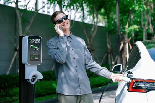 Young man travel with EV electric car charging in green sustainable city outdoor garden in summer shows urban sustainability lifestyle by green clean rechargeable energy of electric vehicle innards