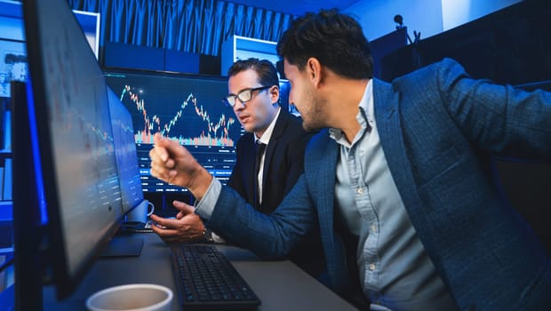 Two stock exchange traders discussing dynamic investment graph in currency rate on monitor at night. Businessman partners coffee meeting in high stock market in neon light at workplace. Sellable.