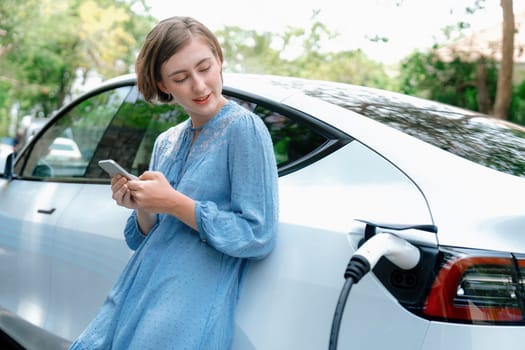 Holiday road trip vacation by the green countryside nature with beautiful young woman checking battery status from smartphone while recharging electric vehicle.Eco-friendly travel wit EV car.Perpetual
