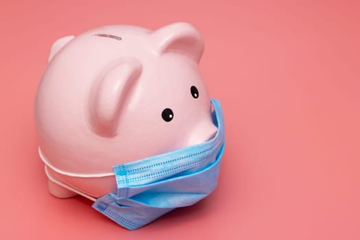 Concept of economic crisis. Piggy bank with protective mask on pink background.