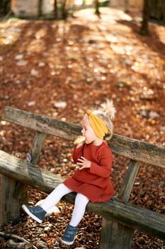 Little girl eating a bun on a wooden bench in the autumn park, dangling her legs. High quality photo