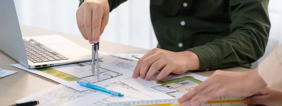 A cropped image of professional engineer using divider to measure blueprint at meeting table with blueprint, laptop and architectural equipment scatter around. Closeup. Delineation.