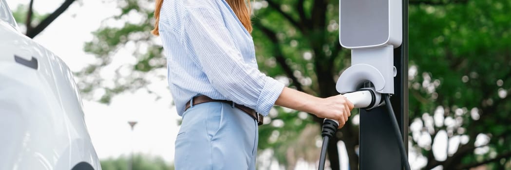 Young woman recharging battery for electric car during road trip travel EV car in natural forest or national park. Eco friendly travel during vacation and holiday. Panorama banner Exalt