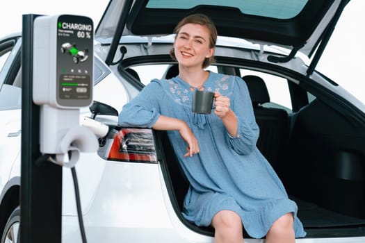 Holiday road trip vacation with young woman sitting in the car trunk with coffee while recharging electric vehicle with alternative energy. Environmental friendly travel wit EV car. Perpetual