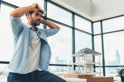 Portrait of skilled architect engineer wear safety helmet while sitting on table with house model, project plan and architectural equipment surrounded by skyscraper view. Civil engineering. Tracery.