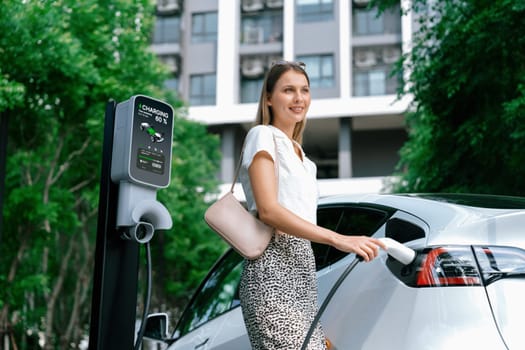 Young woman travel with EV electric car charging in green sustainable city outdoor garden in summer. Urban sustainability lifestyle by green clean rechargeable energy of electric BEV vehicle innards