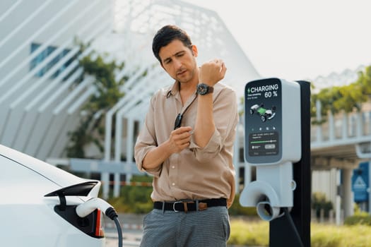 Young man checking time on smartwatch while EV charger to recharging battery from charging station in city mall parking lot. Rechargeable EV car for sustainable eco friendly urban travel. Expedient