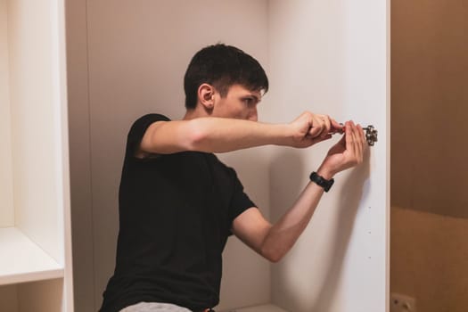 Young handsome caucasian brunette man twists a screw with a screwdriver on a metal fitting while sitting inside a closet in the bedroom, side view close-up with selective focus. Furniture assembly concept.