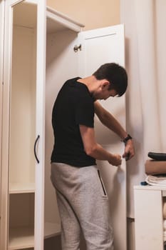 Young caucasian brunette man standing half-side by hand installs black handles on a white wardrobe door, tightening a screw with a screwdriver, close-up bottom view with selective focus. Furniture assembly concept.