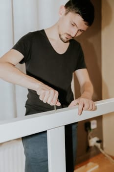 One young brunette Caucasian man screws a screw with his hand into the hole of the white part of a bunk bed, in the evening in the room, close-up side view with selective focus.