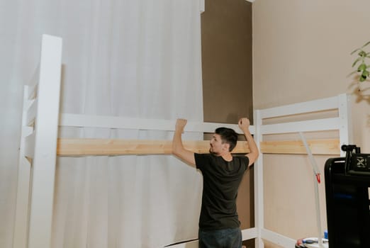 One young brunette Caucasian man stands from the back and connects a board to two white pieces of a bunk bed, in the evening in the room, close-up side view with selective focus.