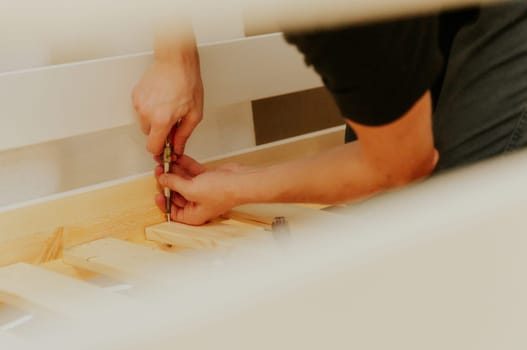 One young unrecognizable Caucasian man sitting sideways on his knees uses a screwdriver to tighten a screw into a bunch of frame boards on a bunk bed, in the evening in the room, top side close-up view with selective focus.