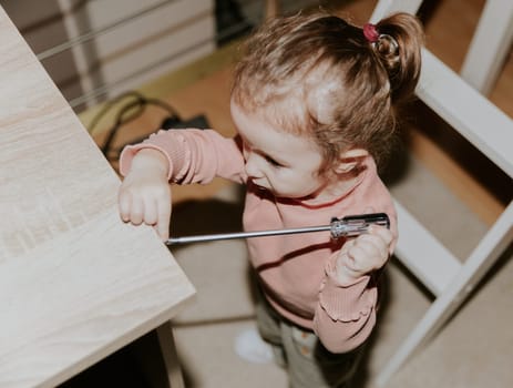 One beautiful little Caucasian girl tightens a screw into an office table with her hands using a screwdriver, standing on the floor in the evening in the room, close-up top view with selective focus.