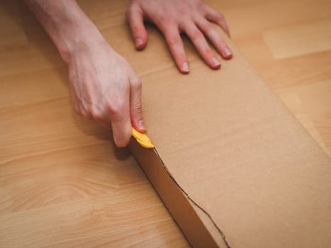 The hands of a caucasian young man cut a long rectangular cardboard box with a yellow construction knife while sitting on the floor in his room, close-up side view. Concept of parcel delivery, furniture assembly, shopping, at home.