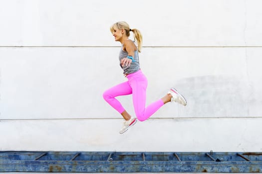 Side view of young female in activewear jumping above ground and smiling while training on street against white wall during fitness workout