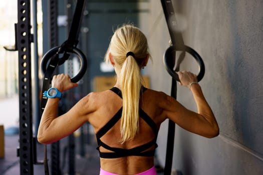 Back view of strong muscular female doing exercises with gymnastic fitness straps rings in modern sport gym club and training alone during workout