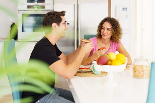 Through blurred green leaves smiling adult couple in conversation, and looking at each other while sitting at dining table in kitchen with fruits in bowl and eating breakfast in daylight at home