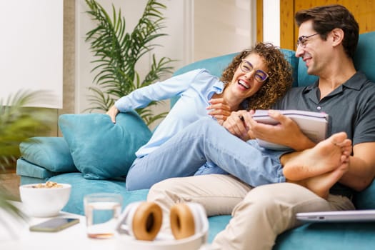 Cheerful young wife in casual clothes sitting with legs on husband, holding notebook while laughing and spending joyful moment together over couch in cozy living room