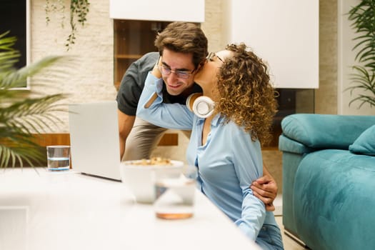 Positive young man and woman with curly brown hair, in smart casual clothes smiling and kissing while looking at laptop screen and working in cozy living room