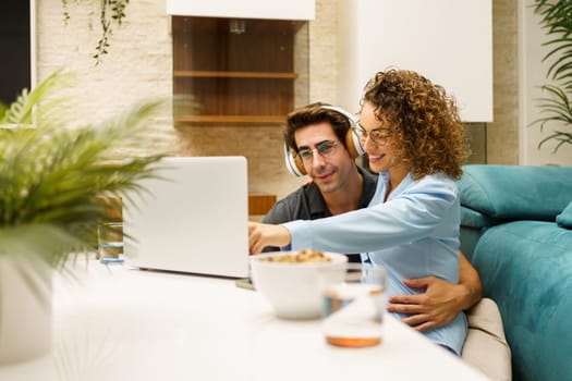 Positive young husband in headphones with curly haired, wife in casual clothes smiling and pointing at screen while sitting together at table with laptop and cups of water in living room