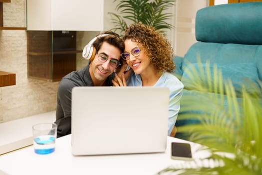Happy young couple in eyeglasses listening to music via wireless headphones while sitting near laptop in living room at home