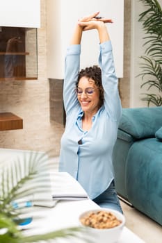 Cheerful young female freelancer in smart casual stretching arms while working on laptop at table in living room at home