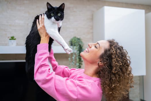 Side view of cheerful young female with curly brown hair, lifting up black and white furred cat looking at camera while playing and bonding at home