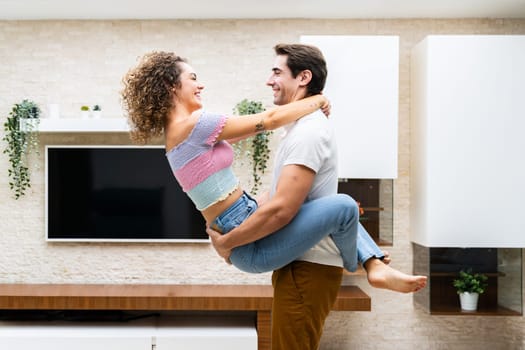 Side view of smiling young couple embracing while having romantic moment in modern apartment against LED TV and looking at each other happily