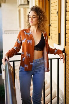 Smiling young female in casual clothes and leaning on balcony of modern residential building while looking away