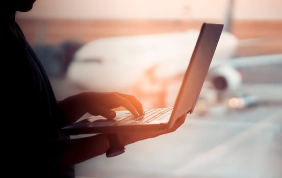 A young man is working on a laptop at the airport while waiting to board the plane. A man is engaged in business, buys tickets, studies and communicates via the Internet at sunset.