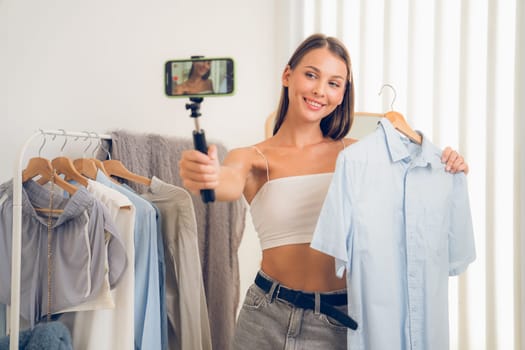 Young woman making fashion video content for social media. Blogger smiles to camera using selfie stick and light ring while making persuasive online clothing sell to audience or followers. Unveiling