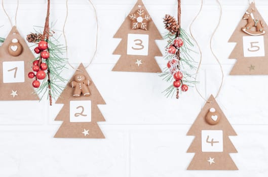 Five handicraft Christmas trees with numbers 1,2,3,4,5, cookies and spruce branches hang on a jute thread on a white brick wall, close-up side view. Advent calendar concept.