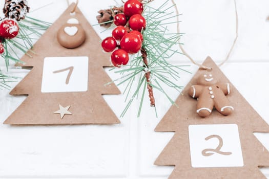Two handicraft trees with numbers 1,2, cookies and a spruce branch hang on a jute thread and on a hanger on a white brick wall, bottom close-up view. Advent calendar concept.