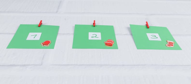 Three green sheets of paper with numbers 1,2,3 and Christmas decor attached with red mini clothespins to a white brick wall, bottom close-up view. DIY advent calendar concept, creativity and minimalism.