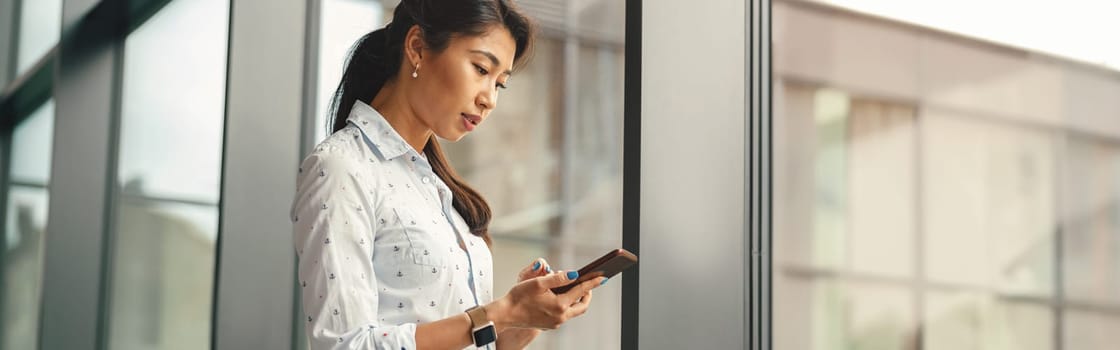 Stylish woman sales manager looking on smartphone standing on modern office background