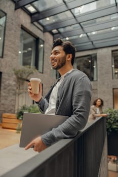 Smiling male freelancer with laptop is standing on coworking background and drinking coffee