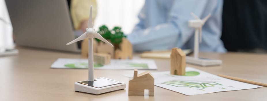 Windmill model represented using clean energy and wooden block represented eco house was scatter around on the table in front of businessman working on laptop. Blurring background. Delineation.