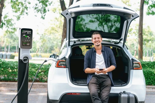 Man using smartphone online banking application to pay for electric car battery charging from EV charging station during vacation holiday road trip at national park or summer forest. Exalt