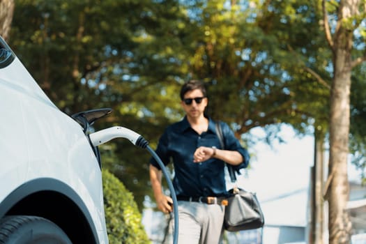 Young man recharge electric car's battery from charging station in outdoor green city park in springtime. Rechargeable EV car for sustainable environmental friendly urban travel lifestyle. Expedient