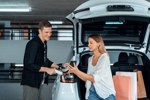 Young couple travel with EV electric car to shopping center parking lot charging in downtown city showing urban sustainability lifestyle by green clean rechargeable energy of electric vehicle innards