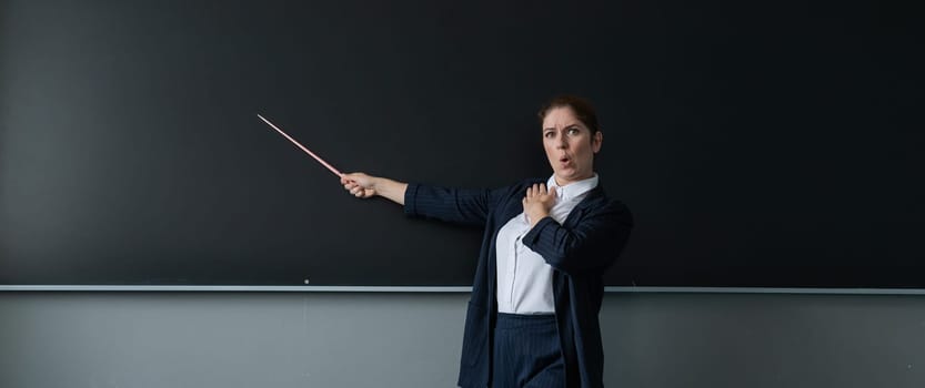 Red-haired caucasian woman in a trouser suit. Thoughtful female teacher with a pointer at the blackboard