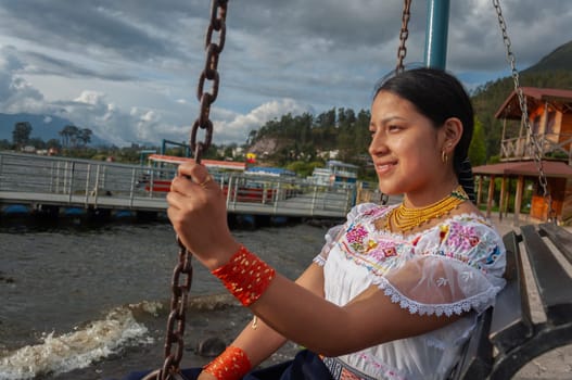 indigenous girl sitting on a swing in front of the dock of a lagoon in Ecuador. The native dresses in her traditional dress and gold jewelry. High quality photo