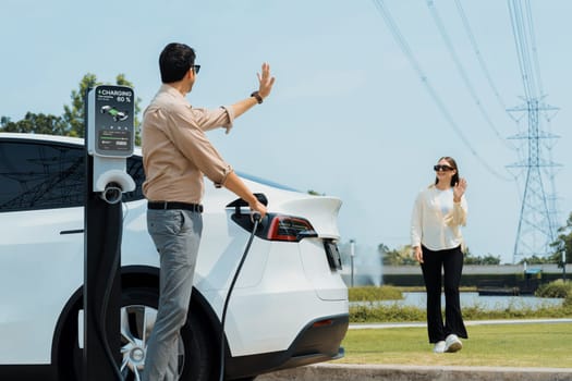 Young couple recharge EV car battery at charging station connected to power grid tower electrical industrial facility as electrical industry for eco friendly vehicle utilization. Expedient