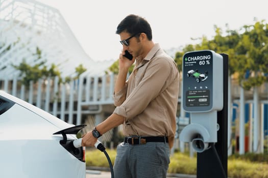 Young man put EV charger to recharge electric car's battery from charging station in city commercial parking lot. Rechargeable EV car for sustainable environmental friendly urban travel. Expedient